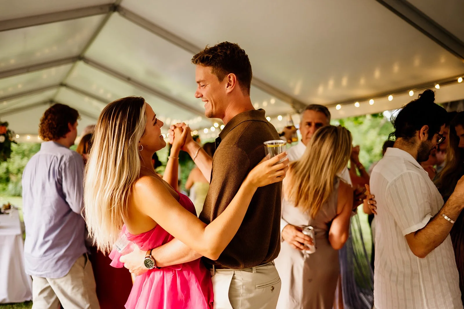 Couple dancing and smiling at a party under a tent, with other people nearby. The woman is in a pink dress, holding a drink, and the man in a brown shirt. String lights decorate the space.