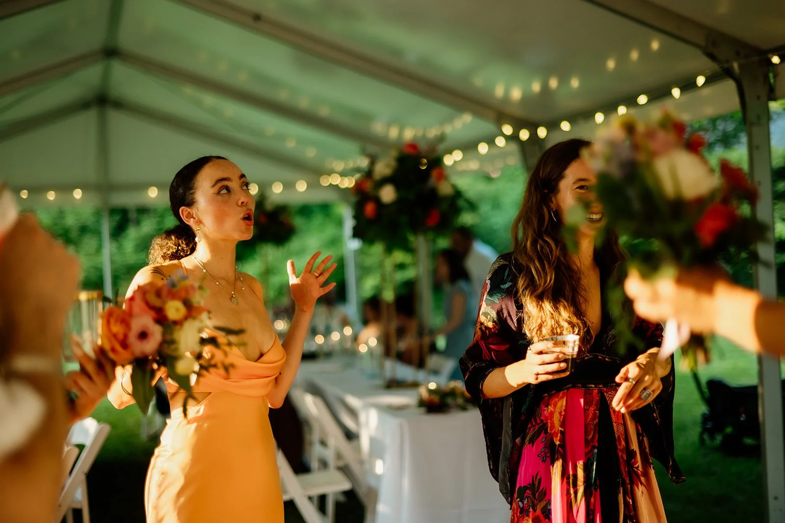 Two women standing under a decorated tent with string lights, one holding a bouquet and the other with a beverage, dressed in formal attire, engaged in conversation during a daytime outdoor event.