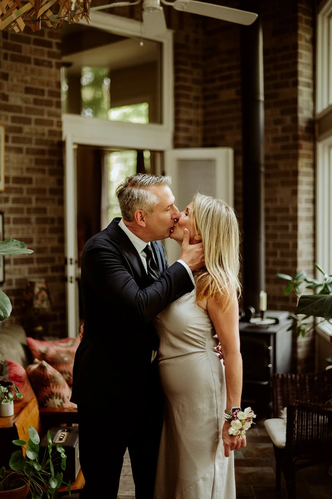 A man in a suit kissing a woman in a white dress in a room with brick walls and plants.
