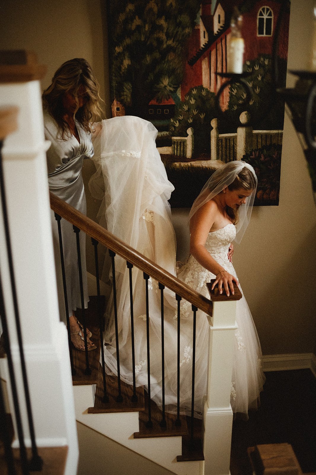 A bride in a wedding dress and veil walking down a staircase inside a house, accompanied by another woman holding part of the dress. A decorative painting hangs on the wall next to them.