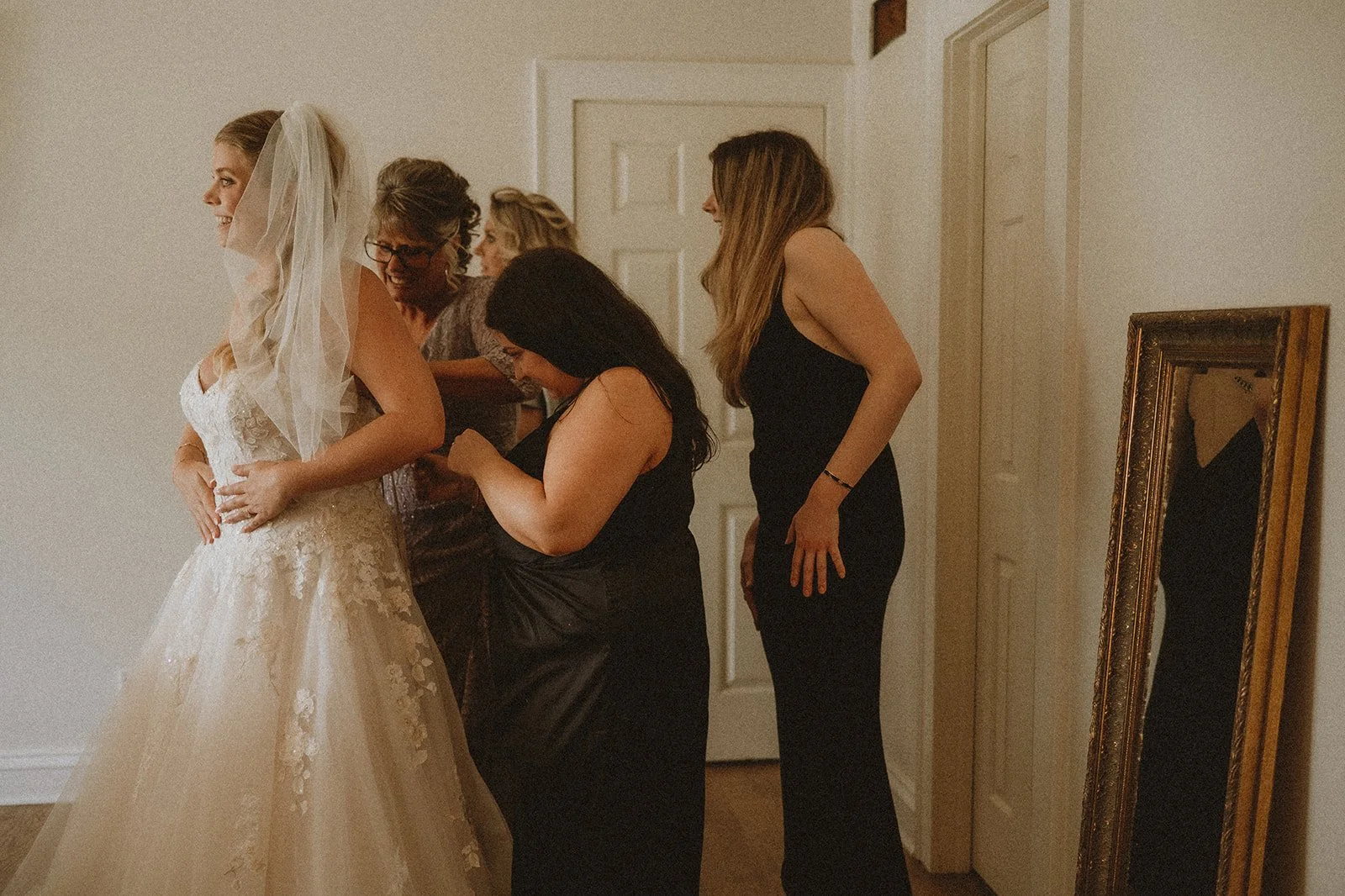 Bride getting ready with help from bridesmaids and a mirror in the background.