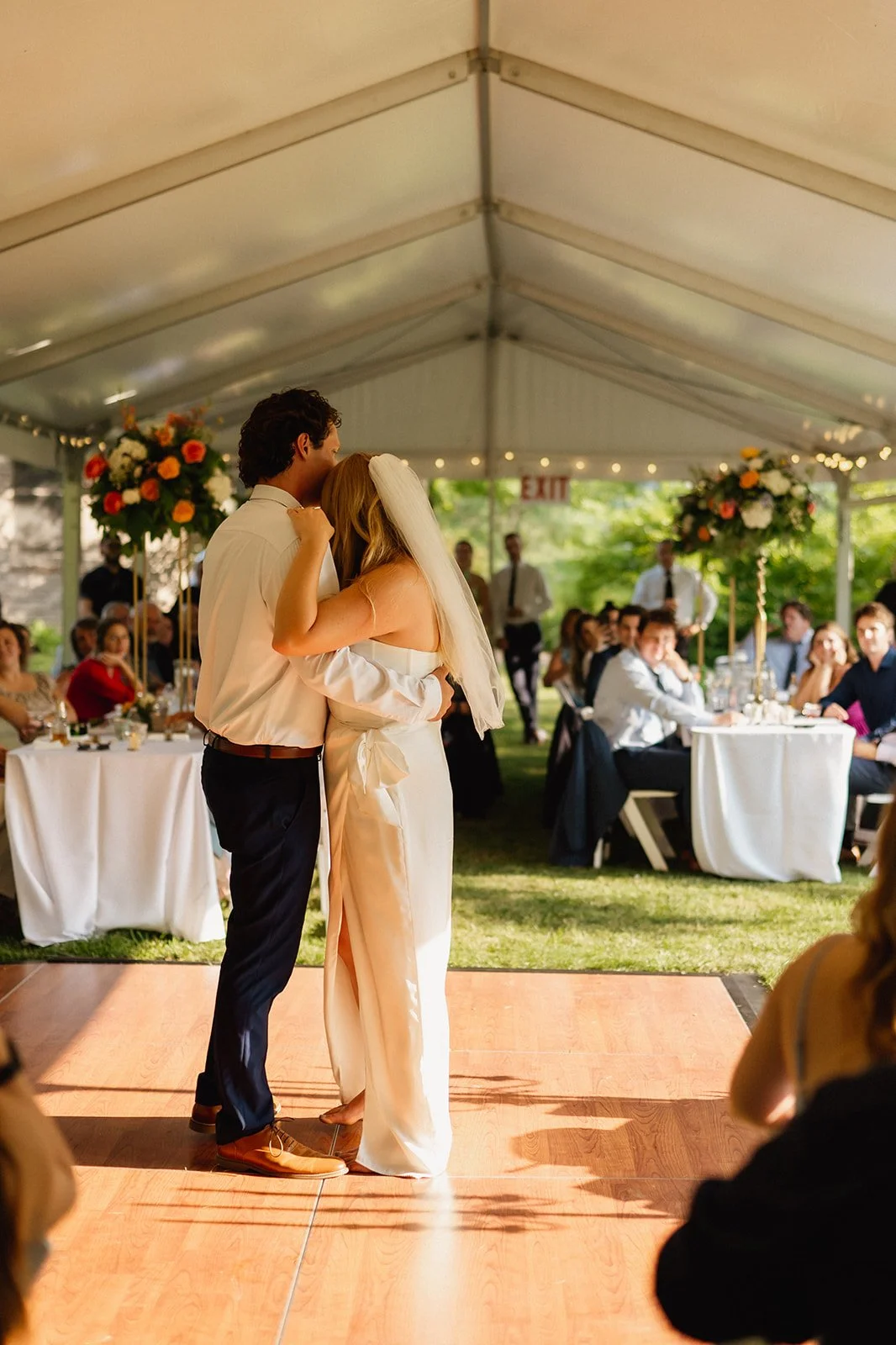 Bride and groom dancing at a wedding reception under a white tent, surrounded by guests seated at tables with floral centerpieces.