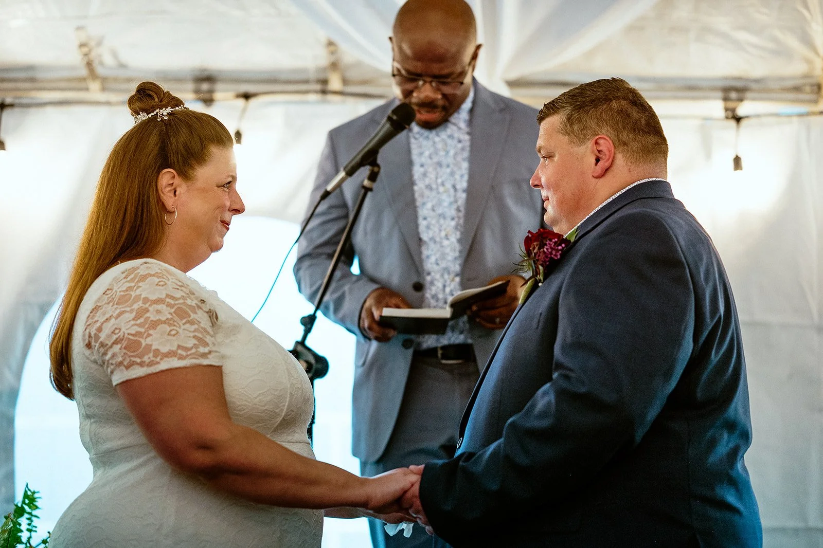 A couple holding hands during a wedding ceremony in a tent, with an officiant reading from a book.