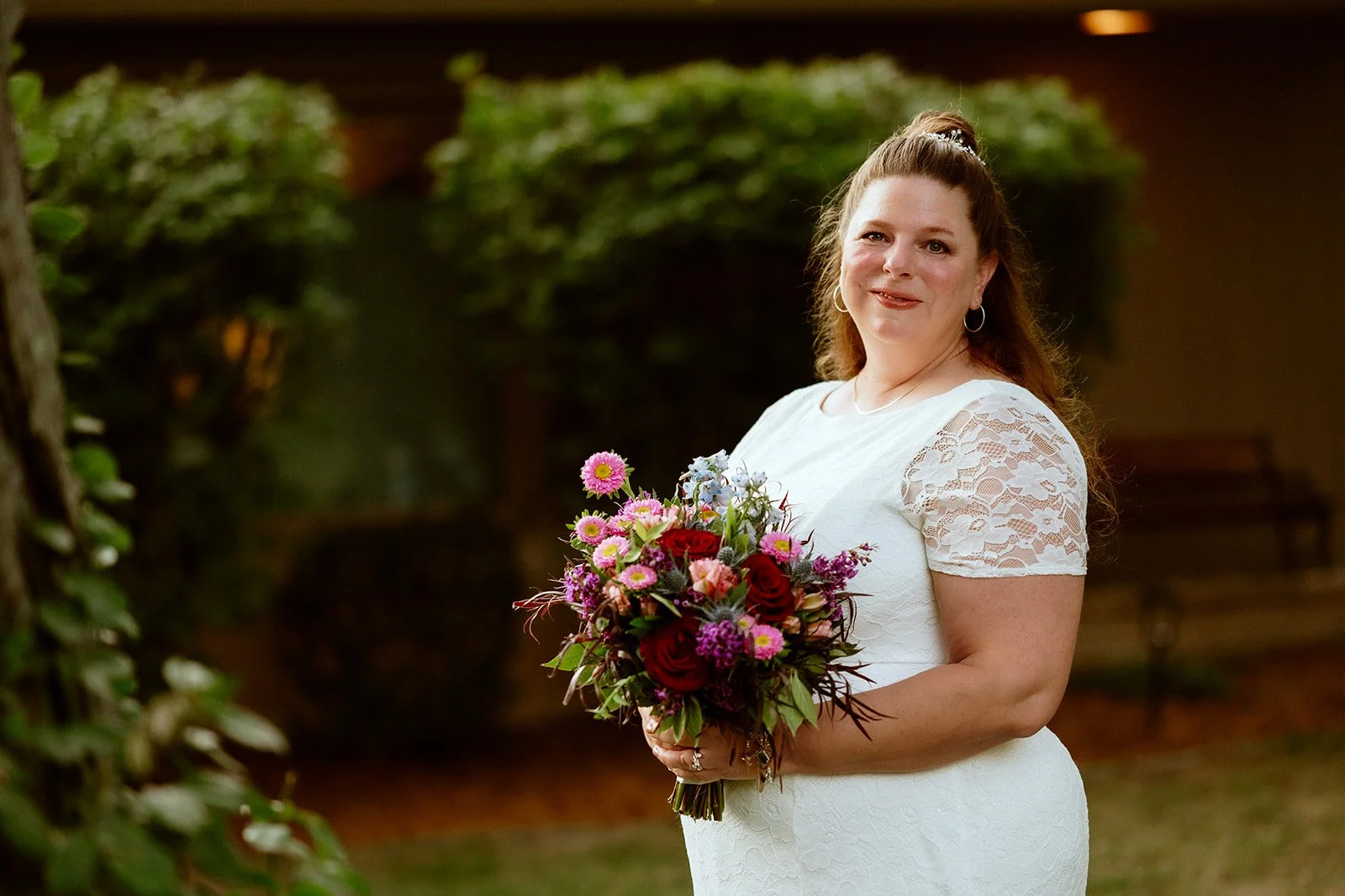 Woman in a white lace dress holding a colorful bouquet of flowers outdoors.