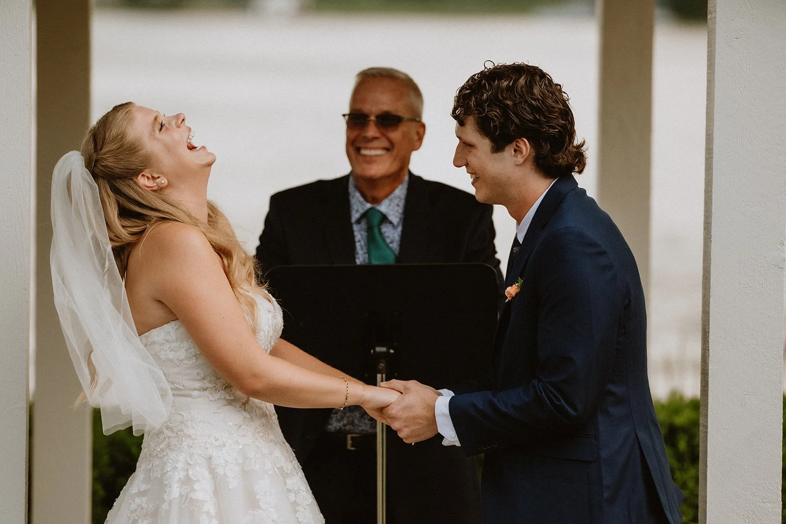 A smiling couple holding hands during a wedding ceremony, with a celebrant in the background under an outdoor structure.