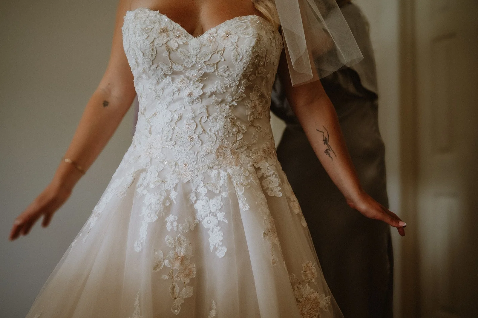 Bride wearing a lace wedding dress with floral embroidery, visible arm tattoo, and veil.
