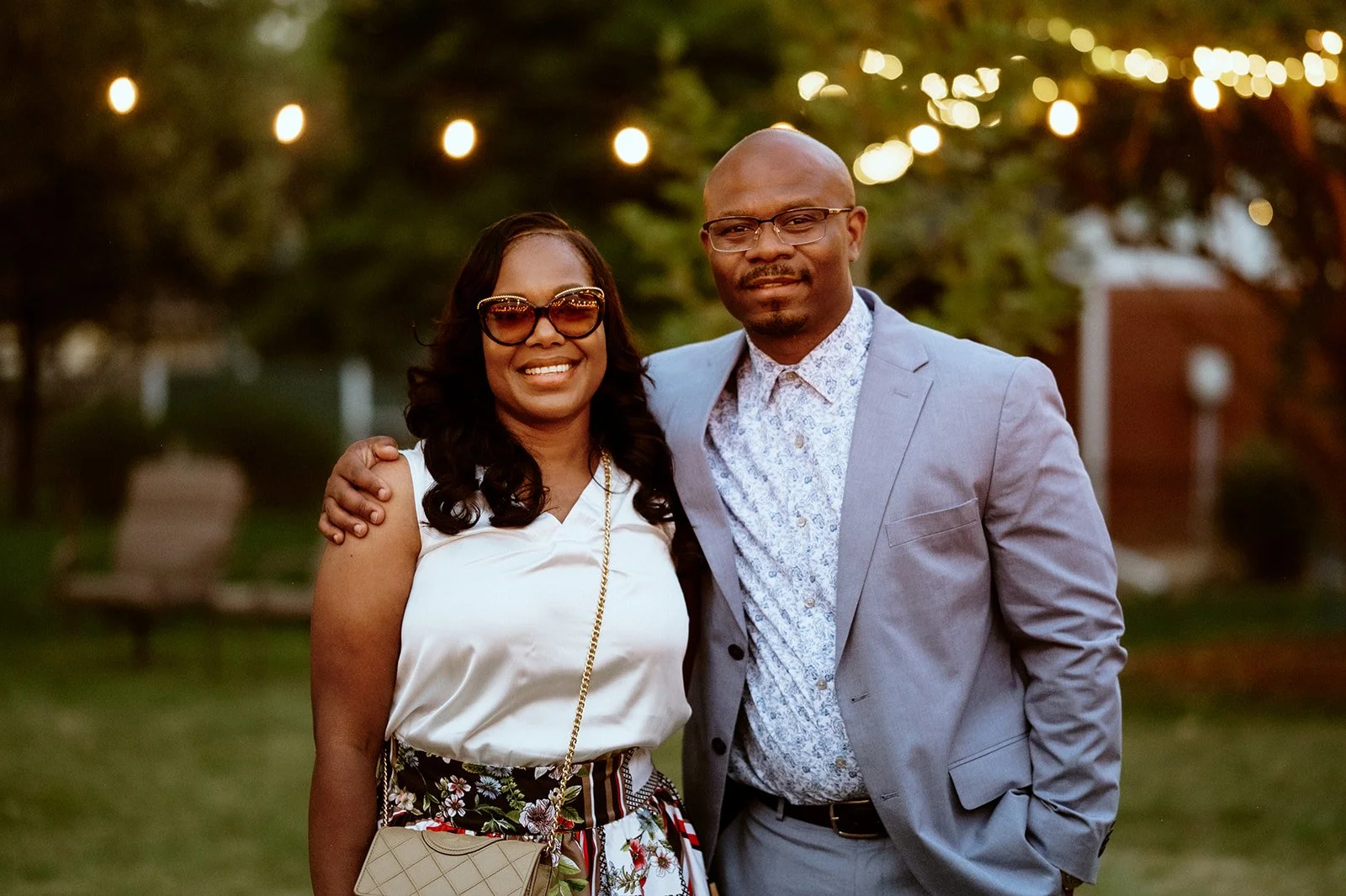 A couple posing outdoors with decorative lights in the background. The woman is wearing sunglasses, a white top, and a floral skirt. The man is wearing a light gray suit and a patterned shirt.