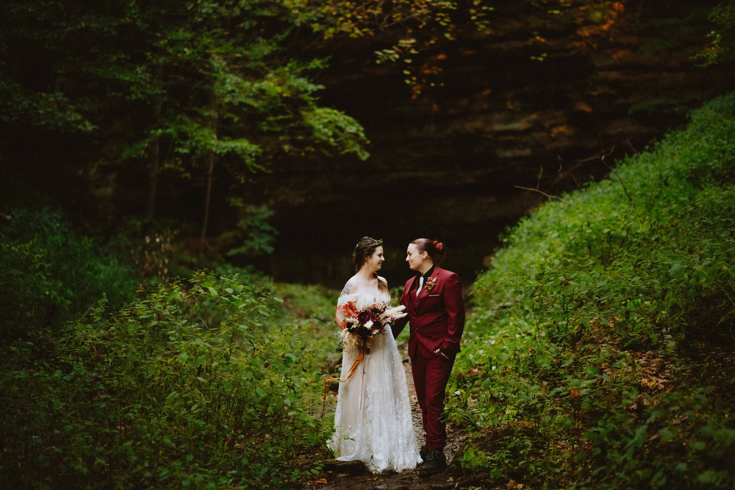 A couple standing on a forest path, one in a wedding dress and the other in a burgundy suit, surrounded by greenery and trees.