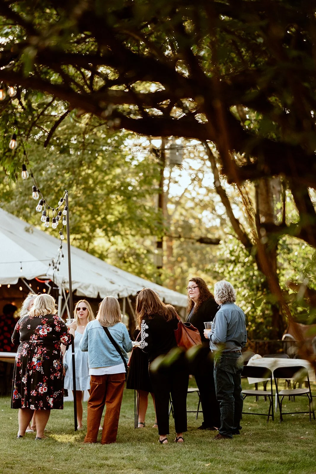 Group of people socializing outdoors at an event with string lights and a large white tent.
