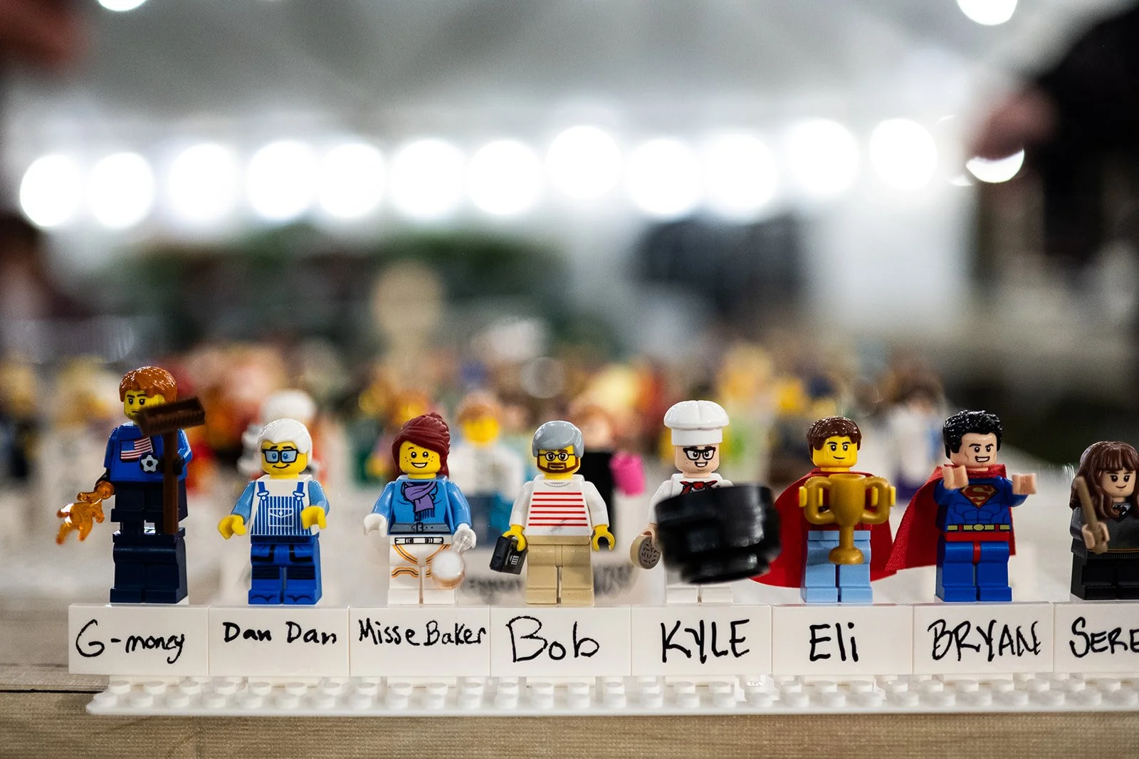 Close-up of several custom LEGO minifigures with name labels on a white base, in a brightly lit room.