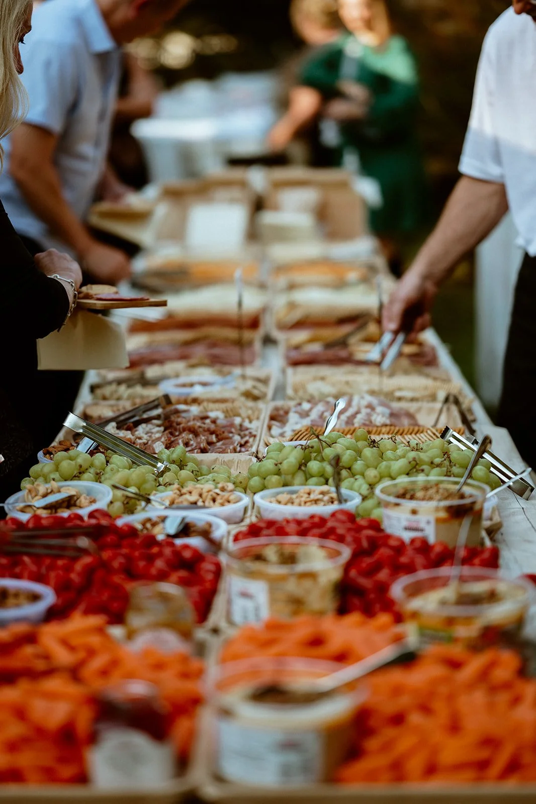 Buffet table with assorted foods including grapes, crackers, nuts, sliced meats, breads, dips, and vegetables, with people serving themselves.