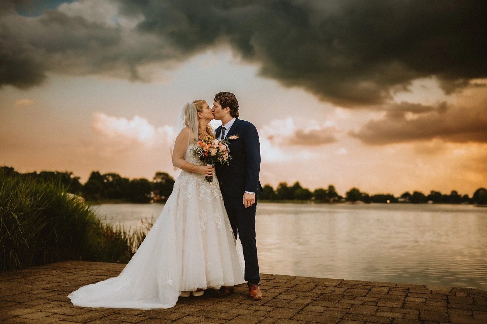 Bride and groom kissing by a lake under dramatic sky