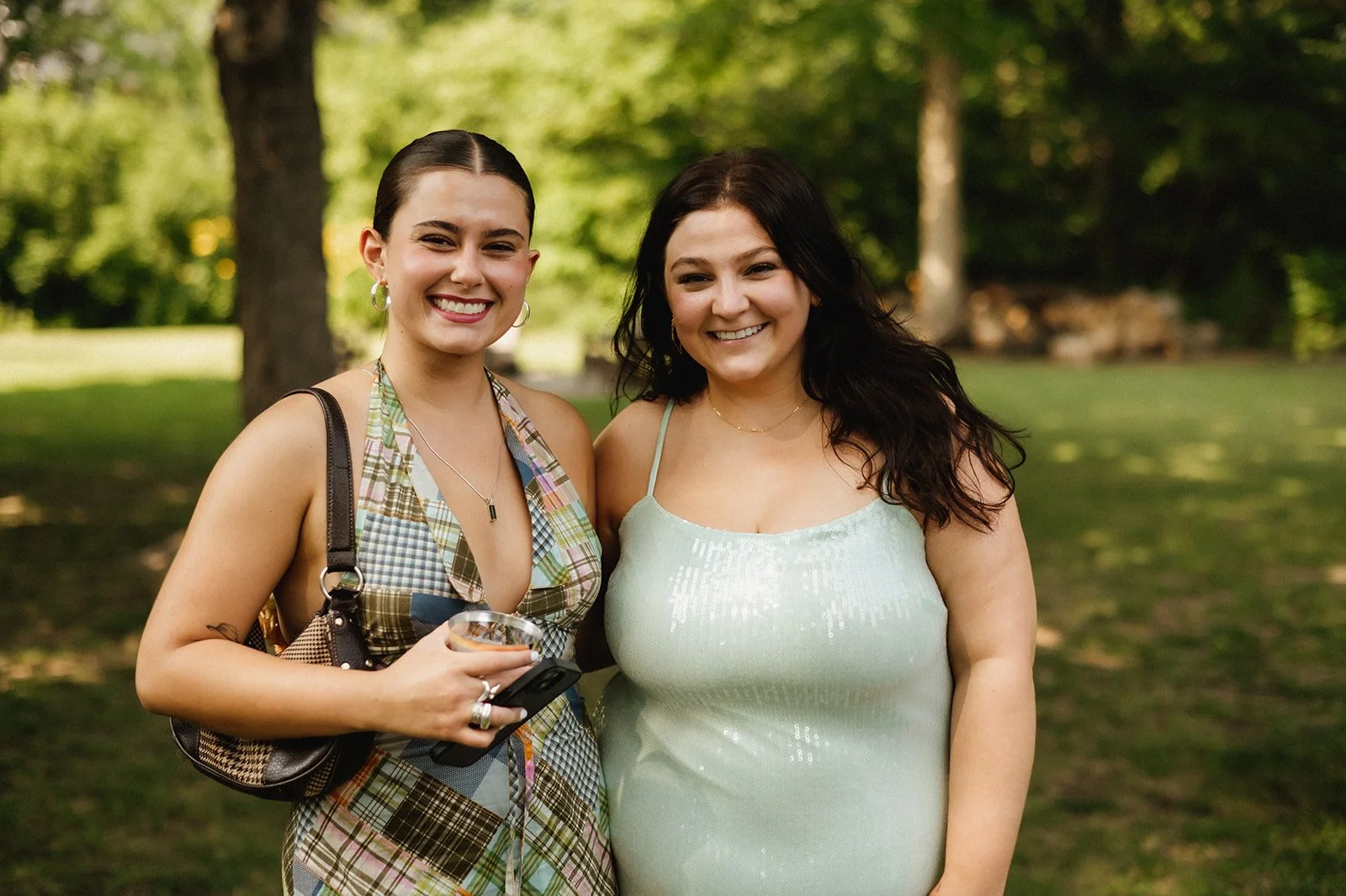 Two women smiling outdoors in a sunny park, one in a plaid dress and the other in a light green sequined dress.