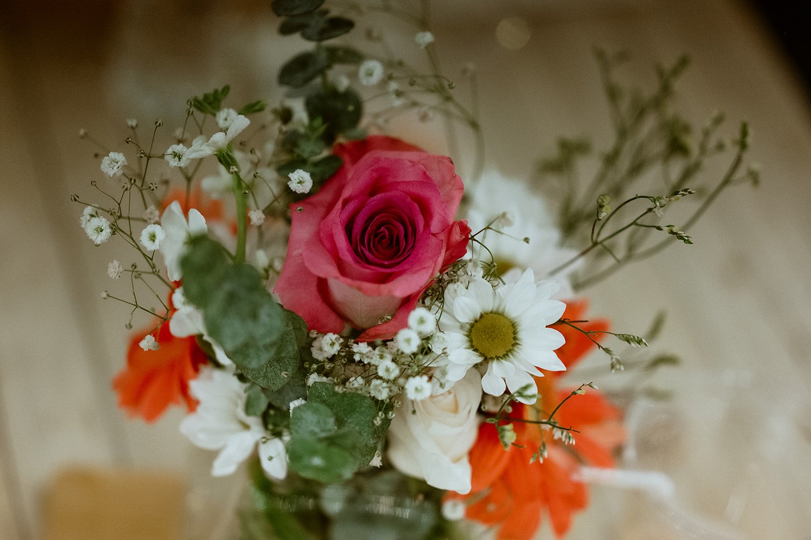 Bouquet with a pink rose, white daisies, orange flowers, and greenery on a wooden background.