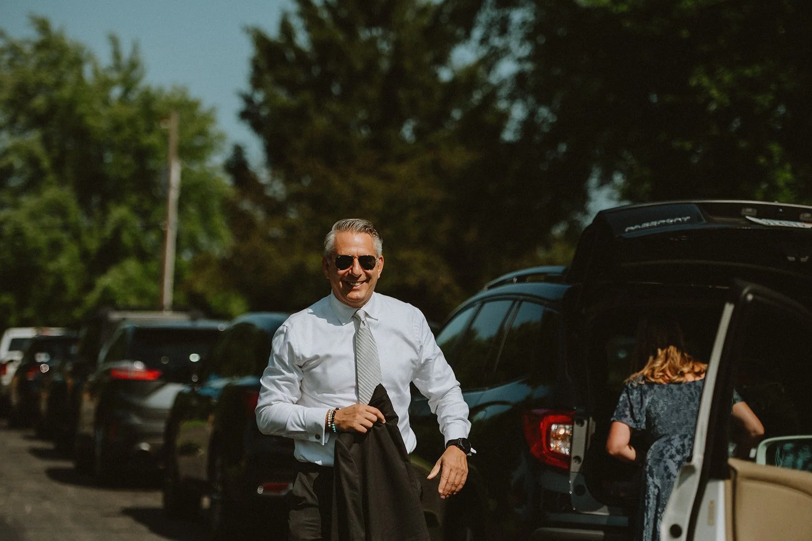 Man in a white shirt and sunglasses smiling near a row of parked cars, holding a jacket. Another person appears near an open car trunk.