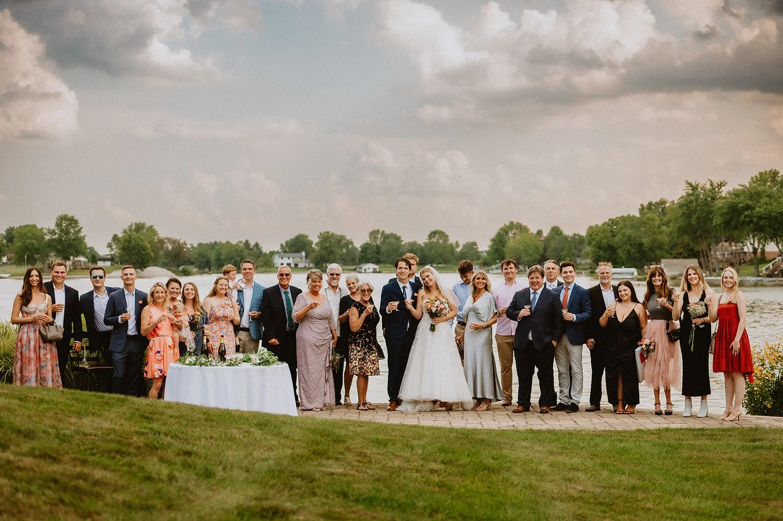 A wedding party posing outdoors by a lake with a table in front and cloudy sky background.