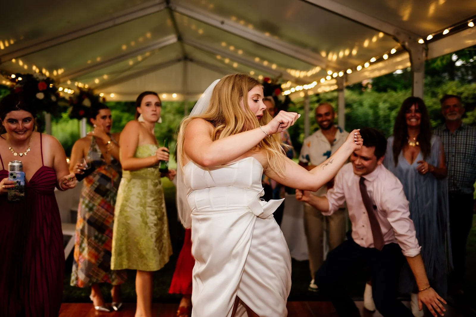 Bride dancing with guests at a wedding reception under a tent with string lights.