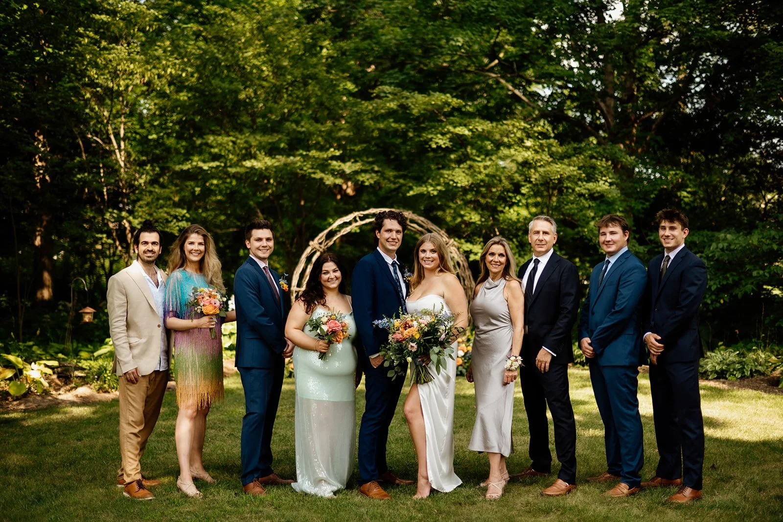 Group of people dressed in formal attire posing outdoors in front of greenery, with two holding bouquets.