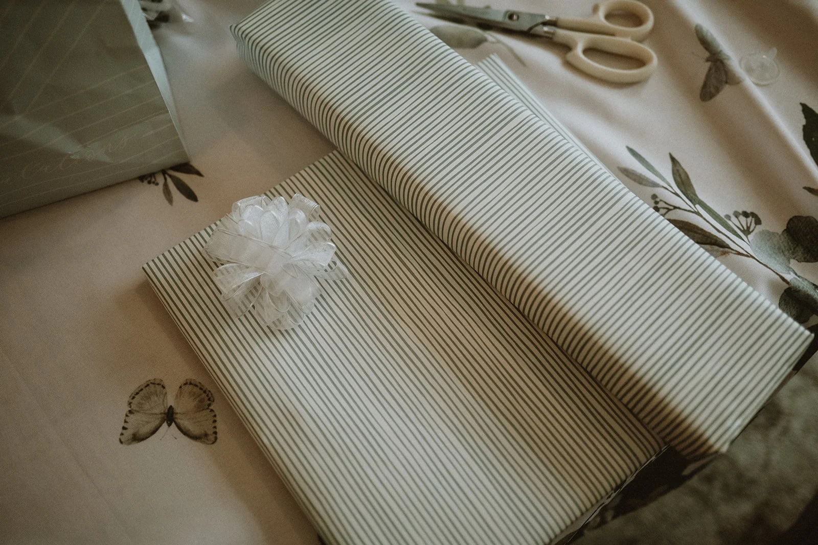 Gift boxes in striped wrapping paper with white bow, scissors, and gift bag on tablecloth with leaf and butterfly patterns.
