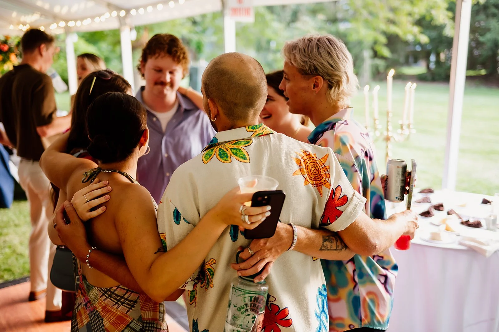 Group of friends gathered outdoors, wearing colorful shirts, sharing a hug during a casual gathering with snacks and drinks on a table nearby.