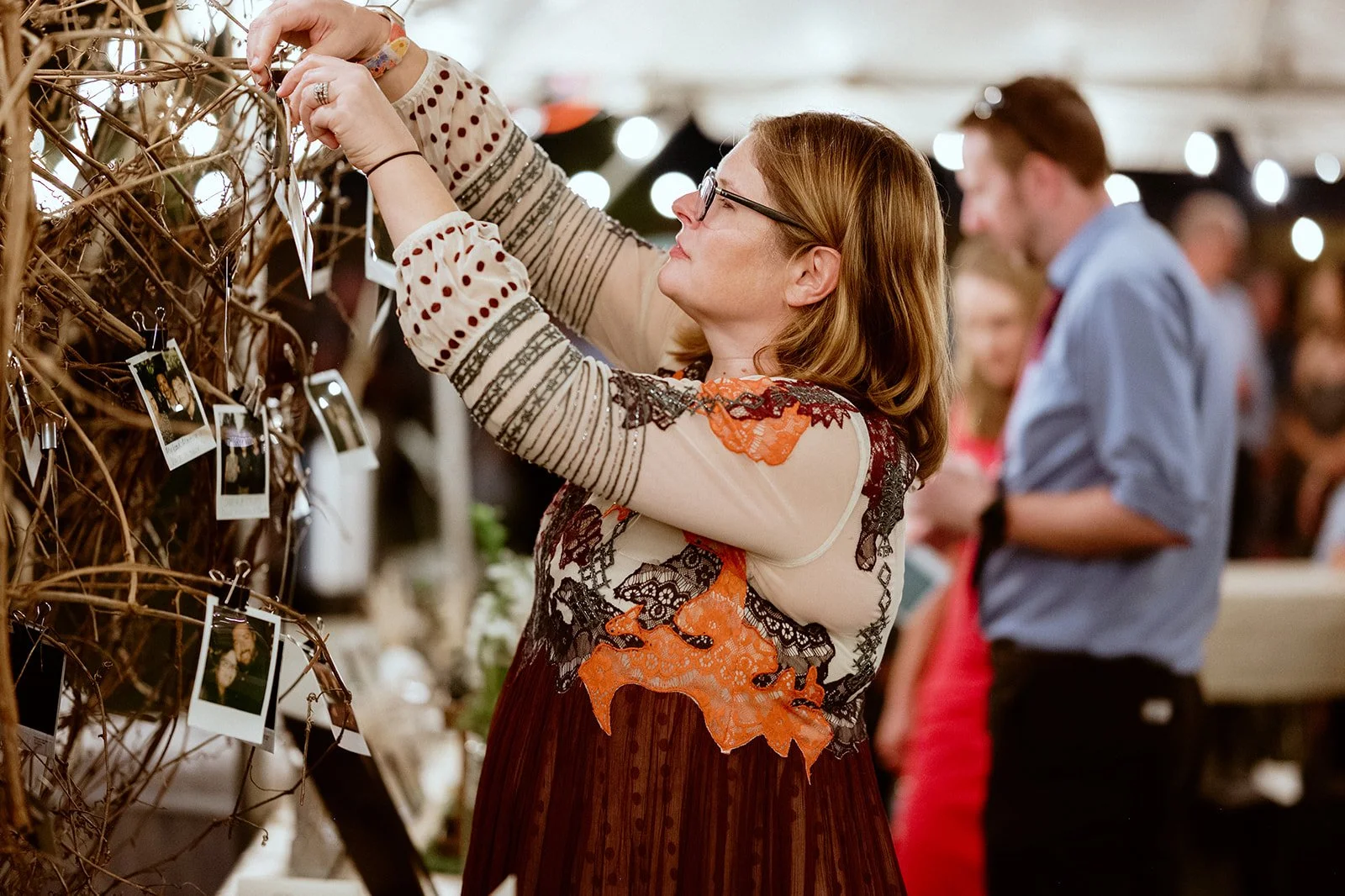 A woman in a colorful dress hanging Polaroid photos on a decorative tree with binder clips at an indoor event, with people in the background.