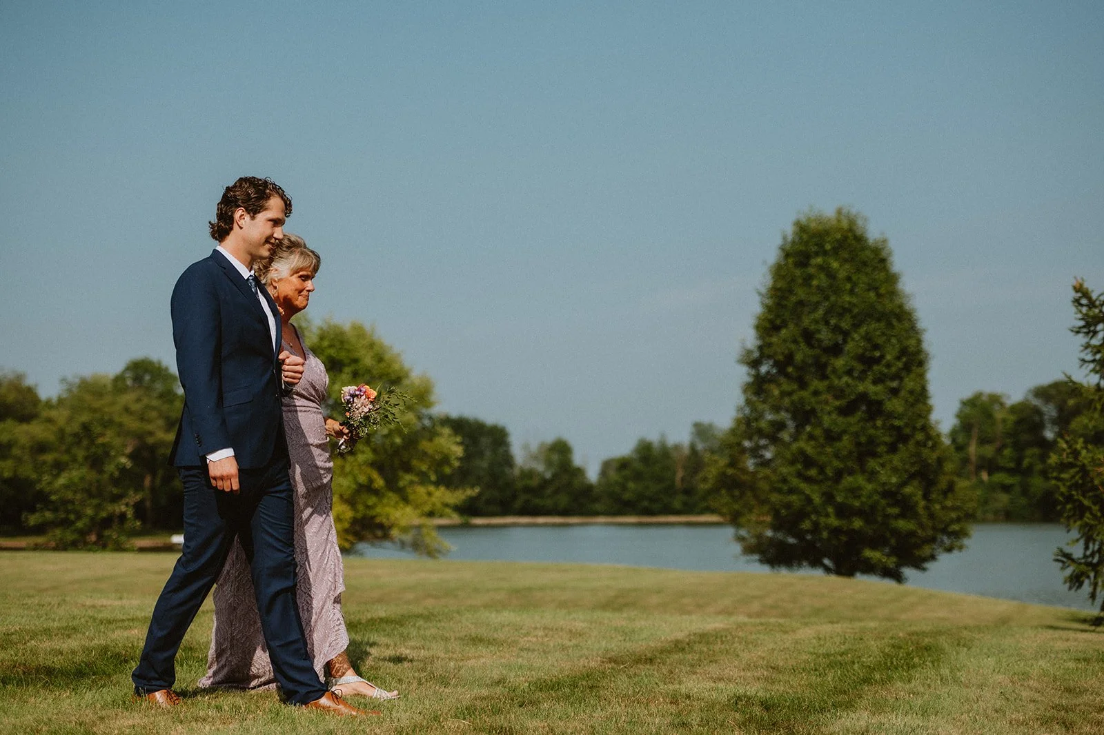 A couple walking on grass near a lake under a clear sky, with trees in the background. The man is dressed in a suit, and the woman is in a formal dress holding a bouquet.