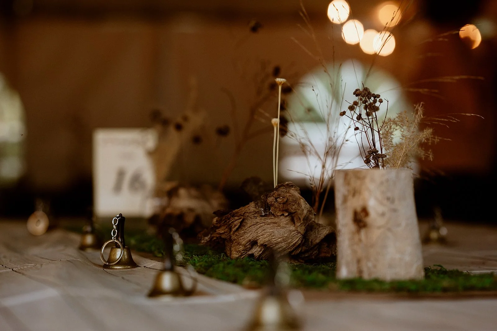 Table centerpiece with dried flowers, wood, moss, small bells, and warm lighting.