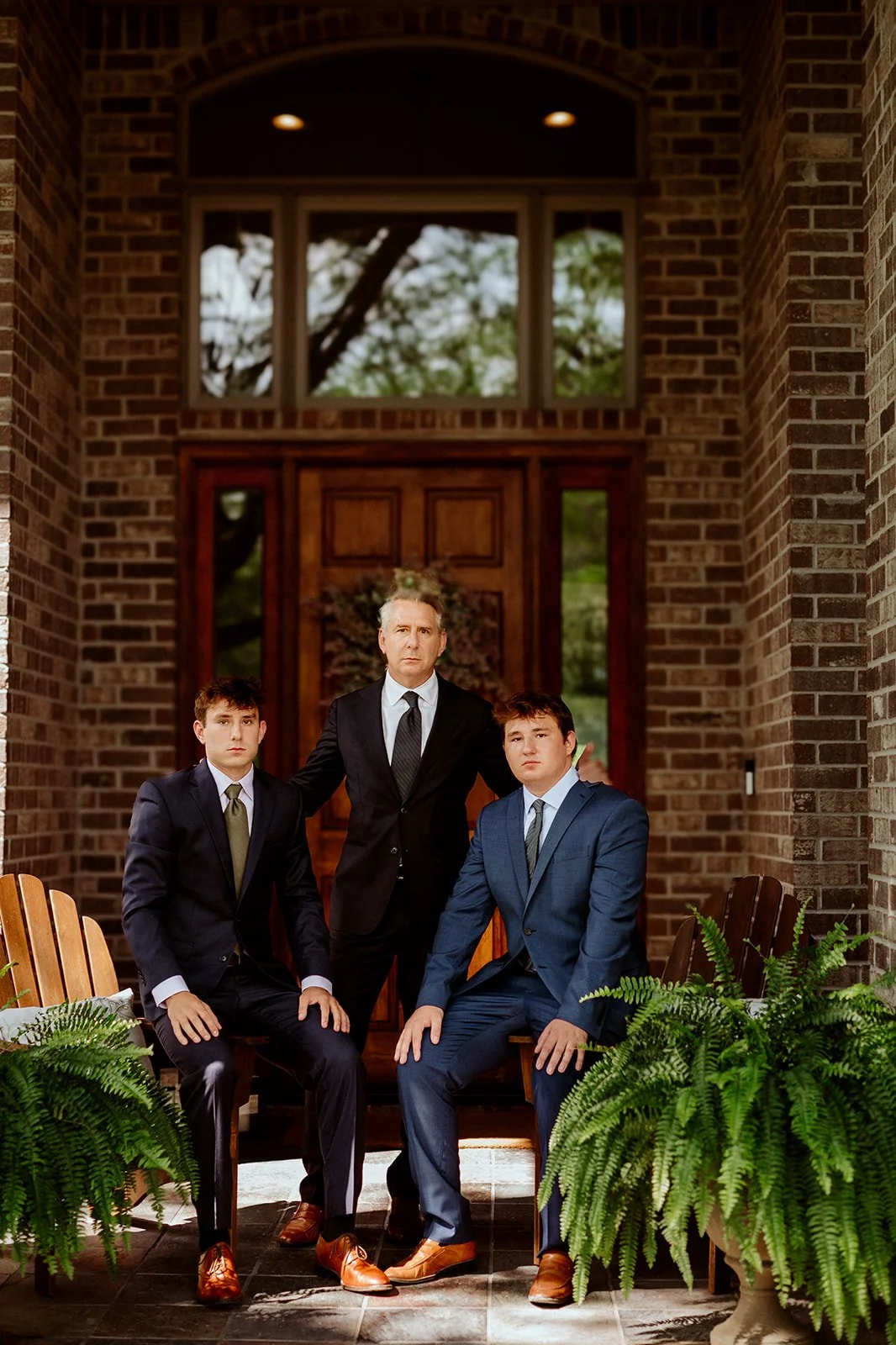 Three men in suits posing on a front porch with brick walls and potted ferns.