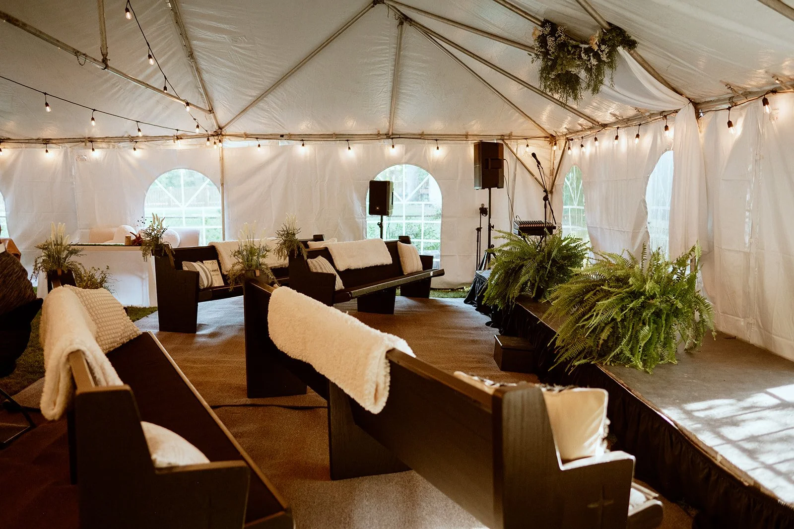 Interior of a tented event space with benches, draped blankets, potted ferns, string lights, and a small stage with a microphone setup.