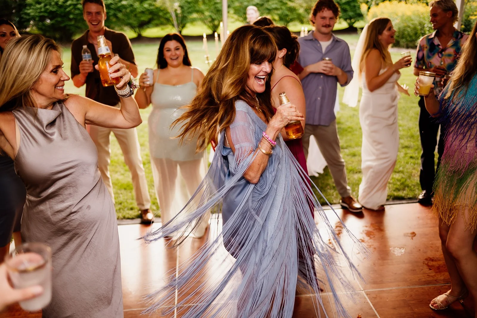 Group of people dancing at an outdoor event, laughing and holding drinks, with a woman in a fringed dress in the foreground.