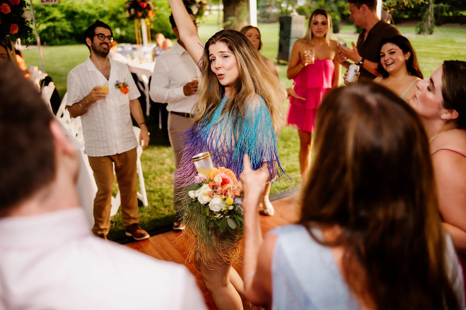 A group of people attending an outdoor party, with a woman in a colorful fringed dress dancing and holding a bouquet. Others around her are smiling and holding drinks.
