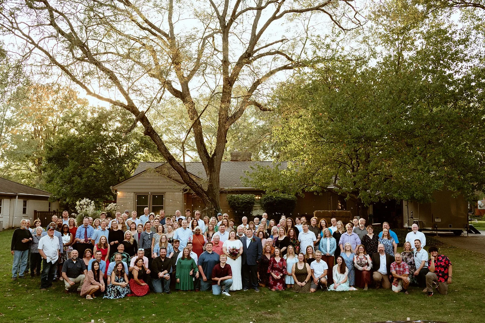 Large group posing outdoors at a wedding, with a bride and groom in the center, in front of a house and trees.