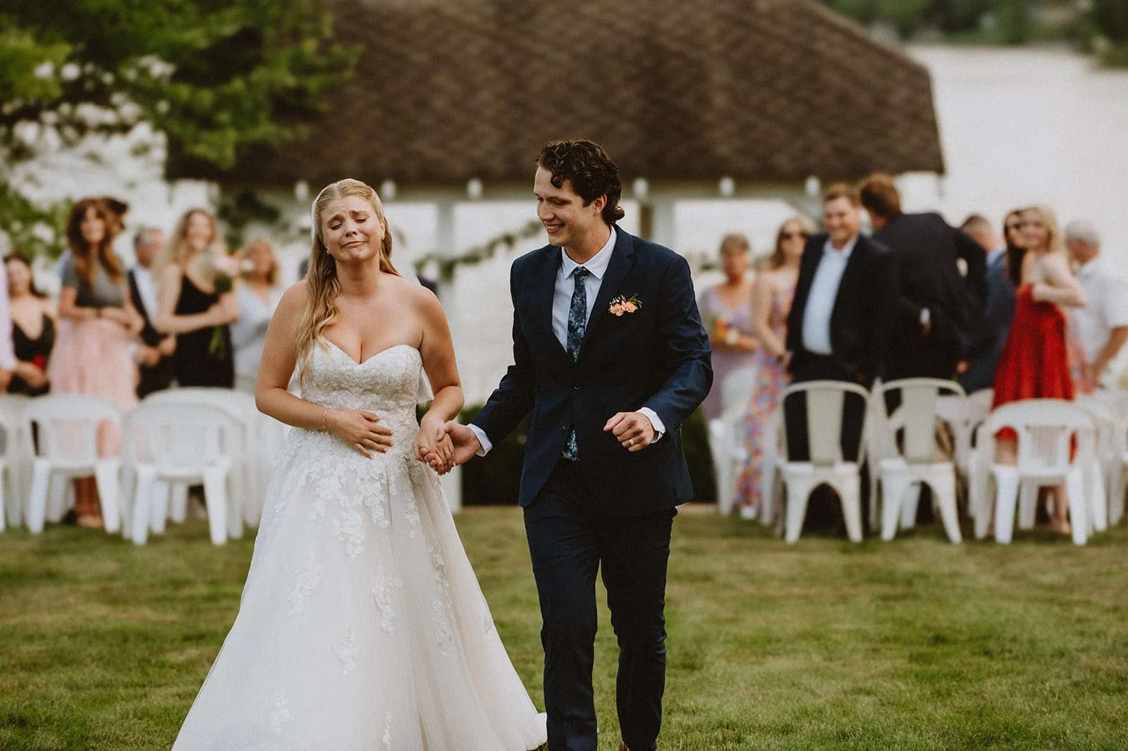 Bride and groom holding hands and walking away from an outdoor wedding ceremony, with guests seated in the background.