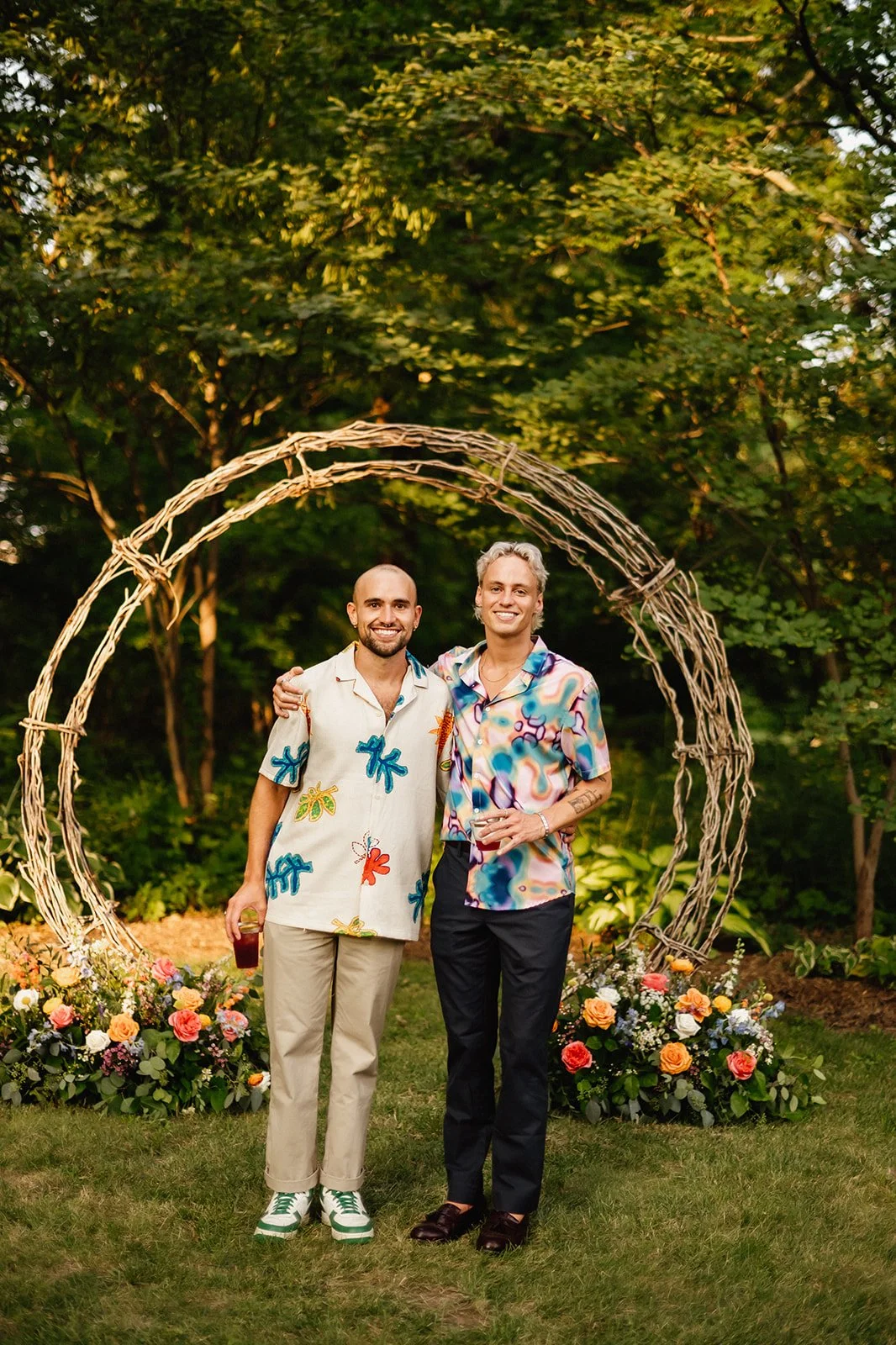 Two men in colorful shirts standing together in a garden setting with flowers and a decorative arch made of twigs.