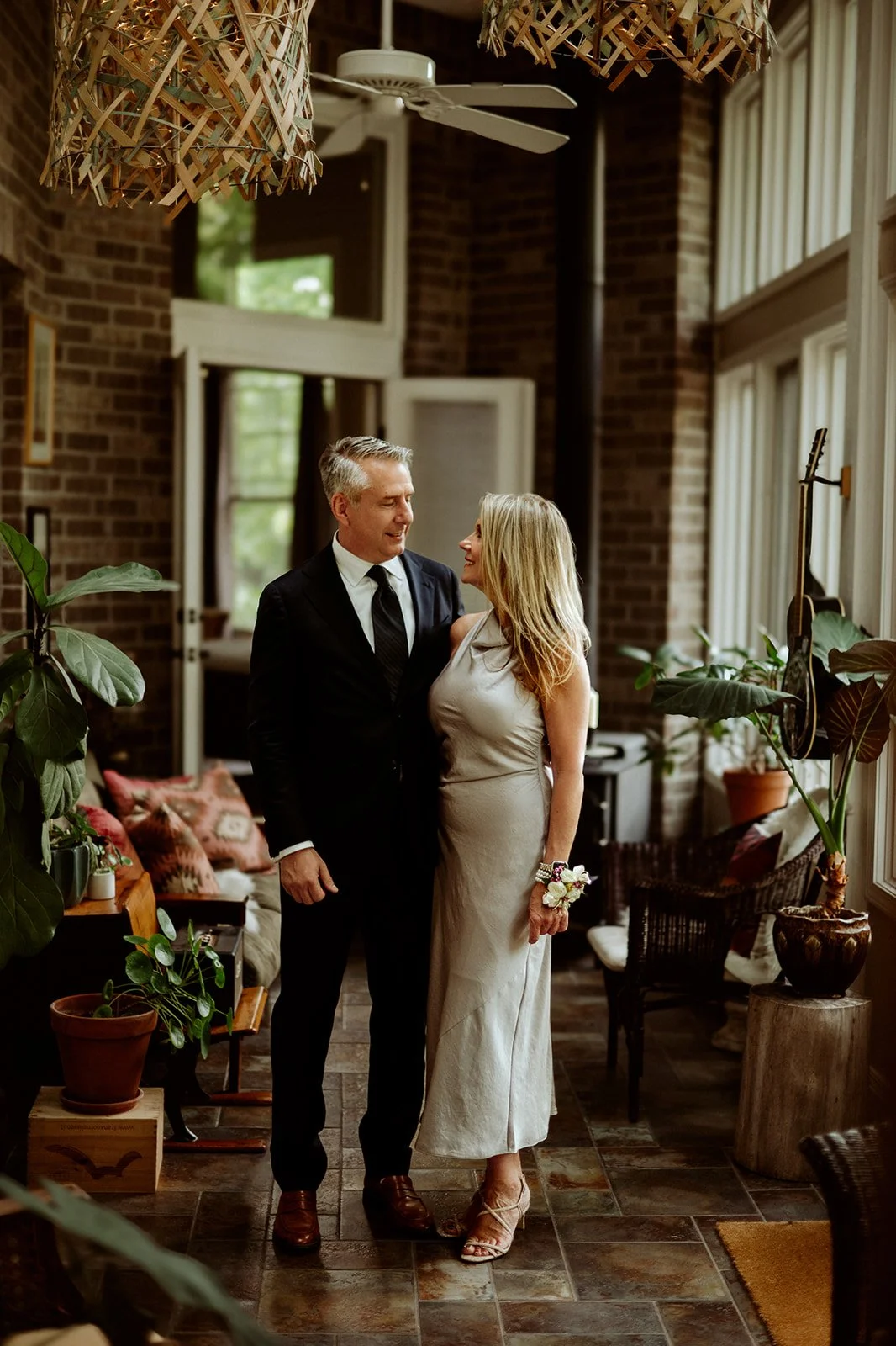 A couple dressed elegantly, standing closely in a stylish, plant-filled room with a guitar hanging on the wall.