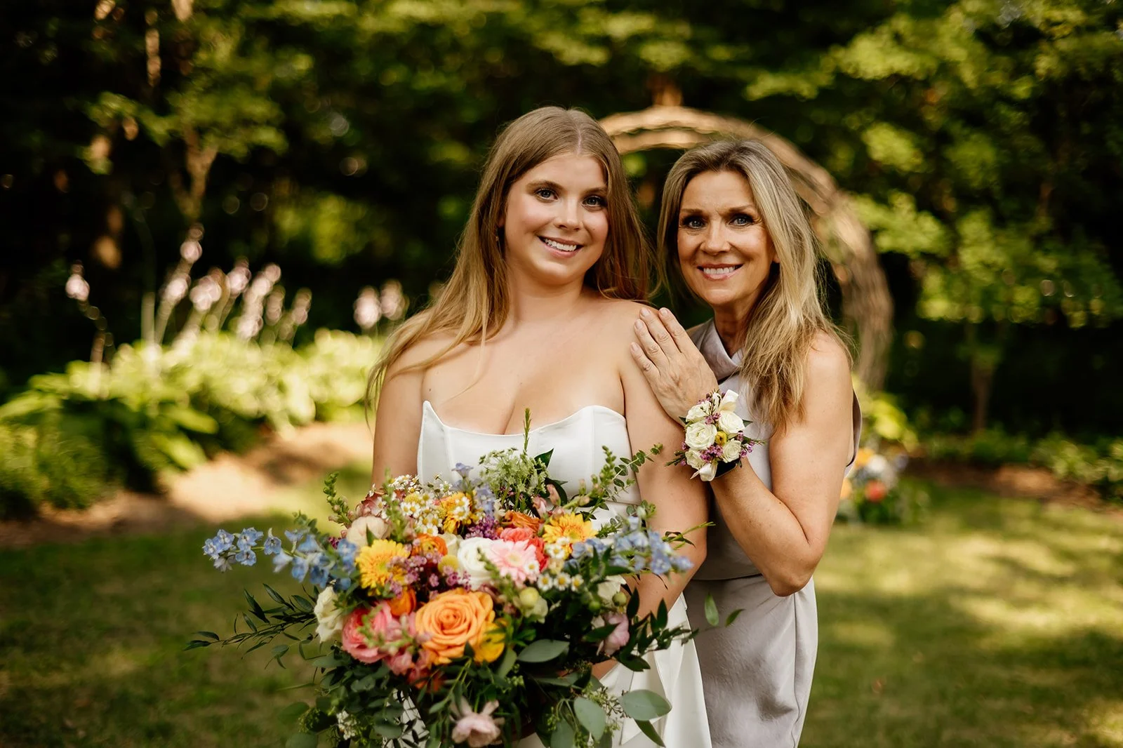 A woman in a white dress holding a colorful bouquet, standing next to another woman in a light-colored dress. They are outdoors in a garden, smiling at the camera.