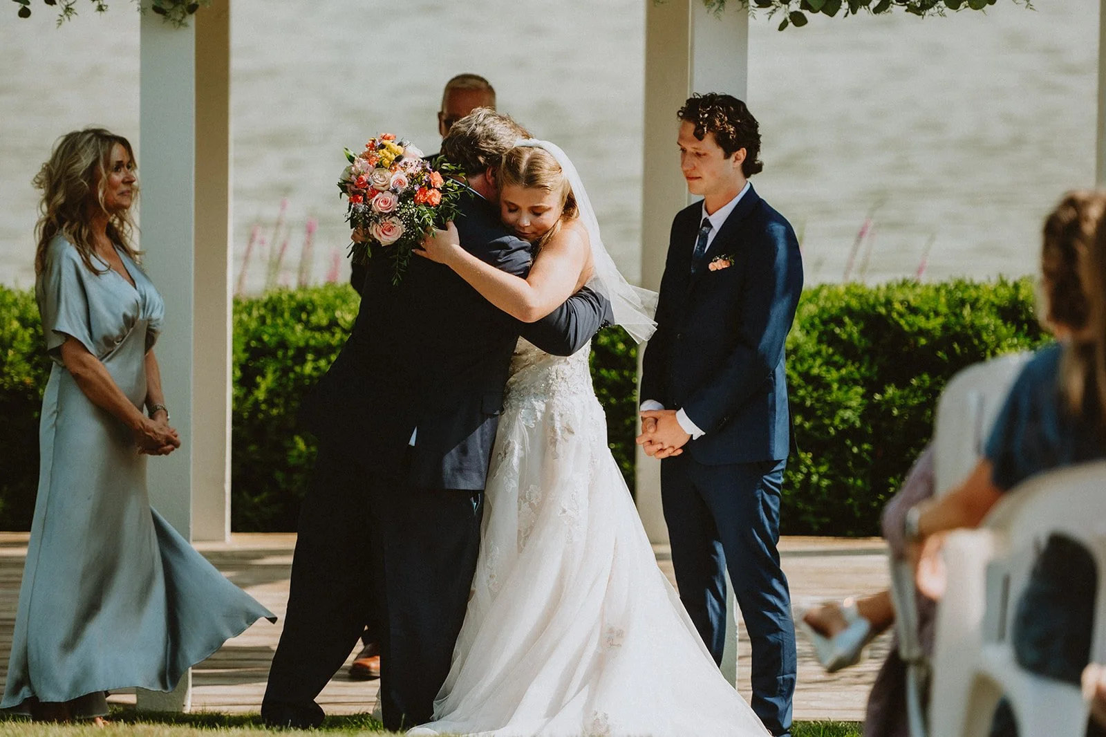 Bride hugging an older man at a wedding ceremony with groom and other guests watching.