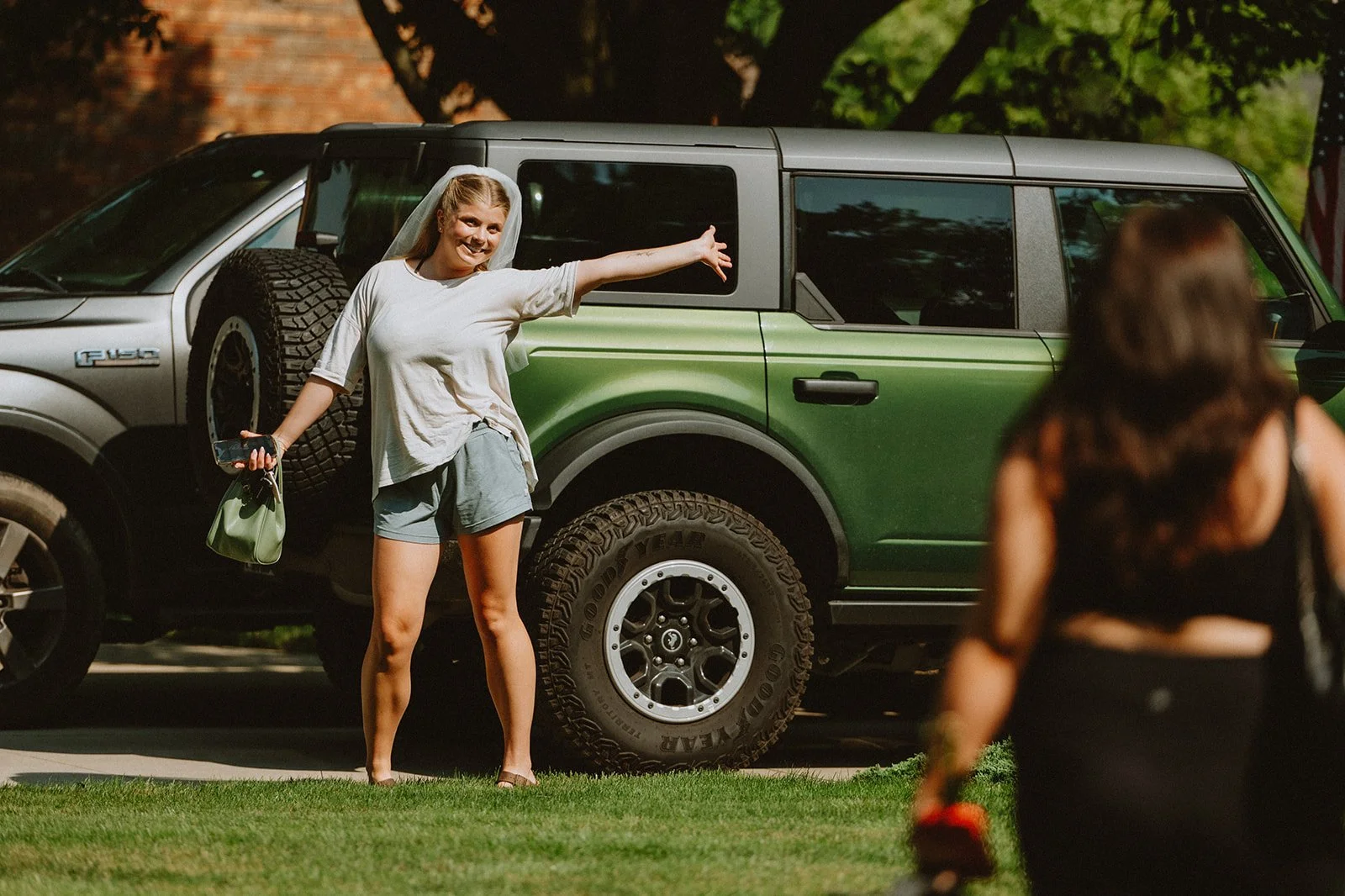 A person posing happily in front of a green Ford Bronco with outstretched arms, wearing casual shorts and a t-shirt. Another person approaches in the foreground, partially visible. The setting is outdoors with sunlight and trees.