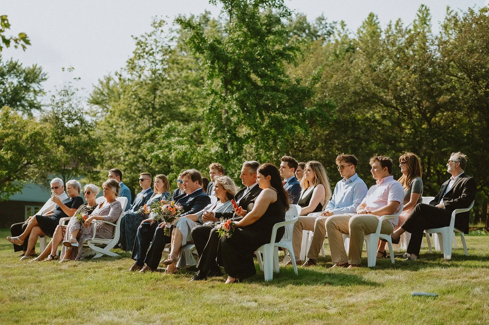 People sitting in chairs outside during a daytime event, possibly a wedding, with trees in the background.
