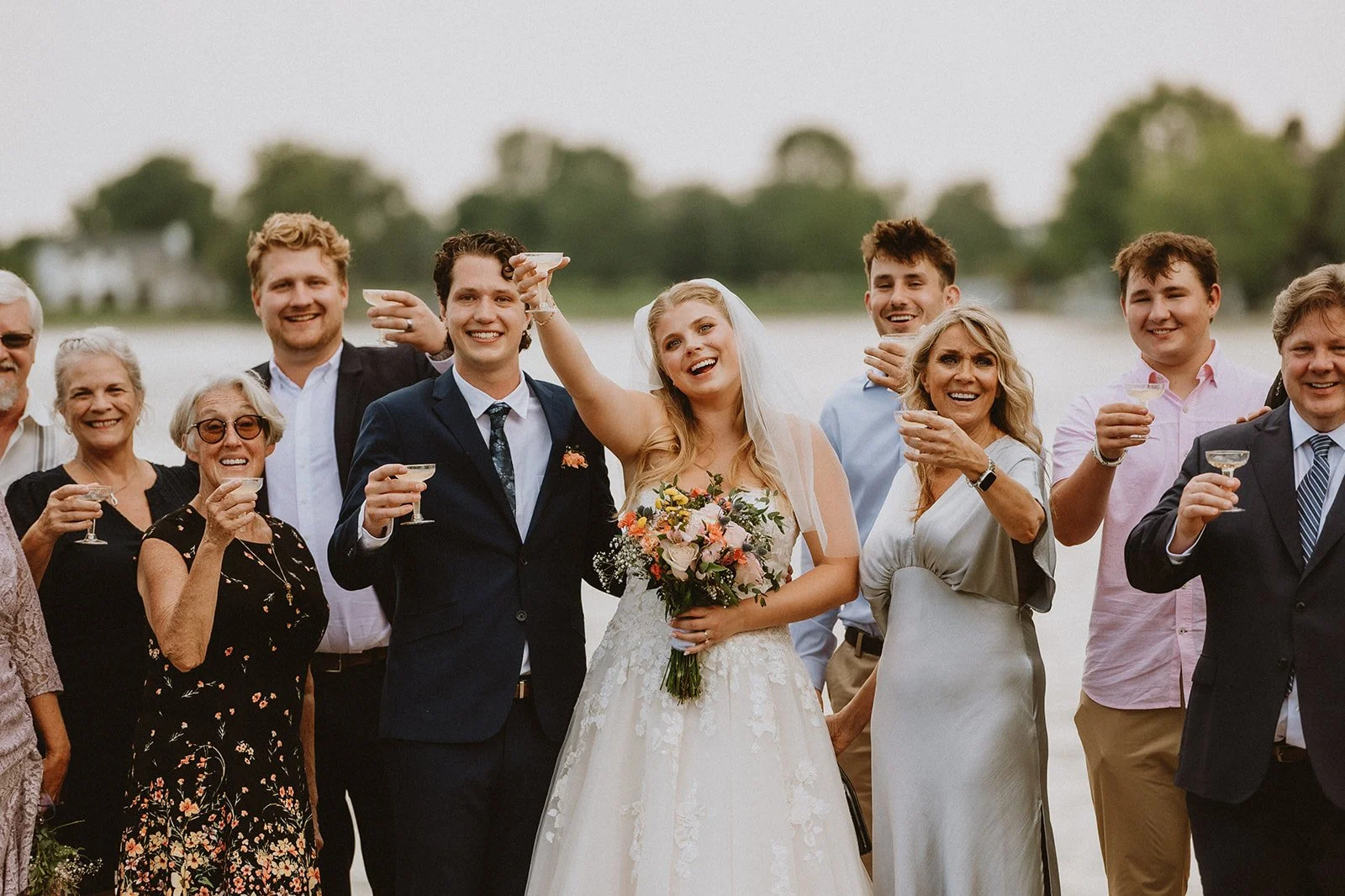 A group of people celebrating at a wedding, with the bride holding a bouquet and others raising glasses for a toast. They are outside with greenery in the background.