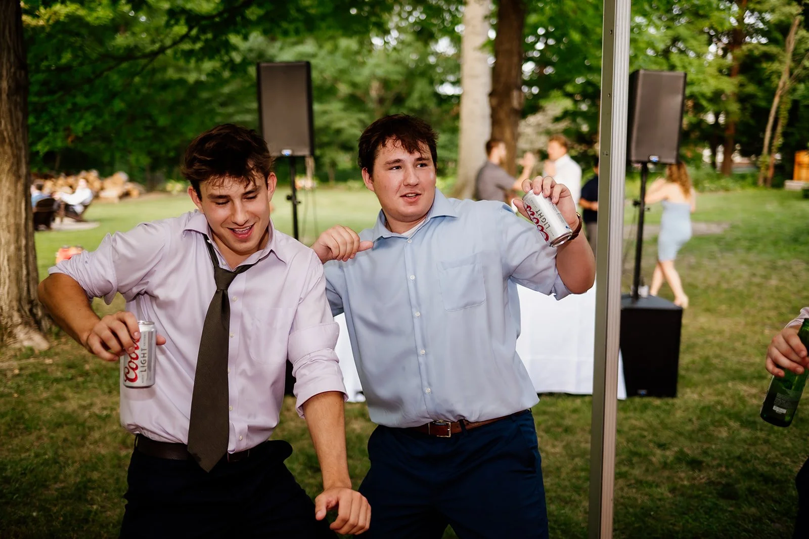 Two young men dancing outdoors at a casual event, each holding a can of Coors Light, with trees and people in the background.