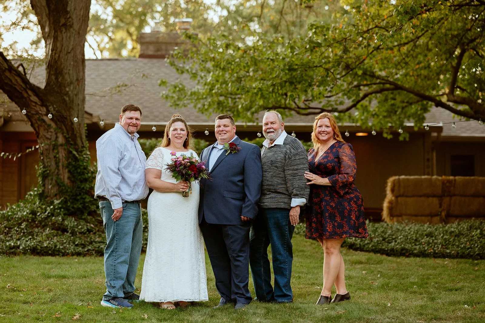 Wedding group photo with bride in a white dress and groom in a suit, surrounded by three other people in casual attire, standing outdoors with a house and trees in the background.