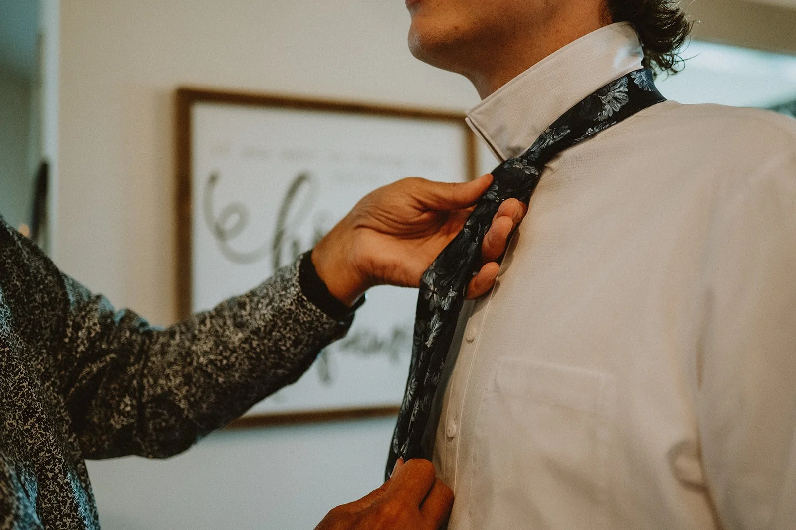 Close-up of a person helping another person adjust a floral necktie over a white shirt.