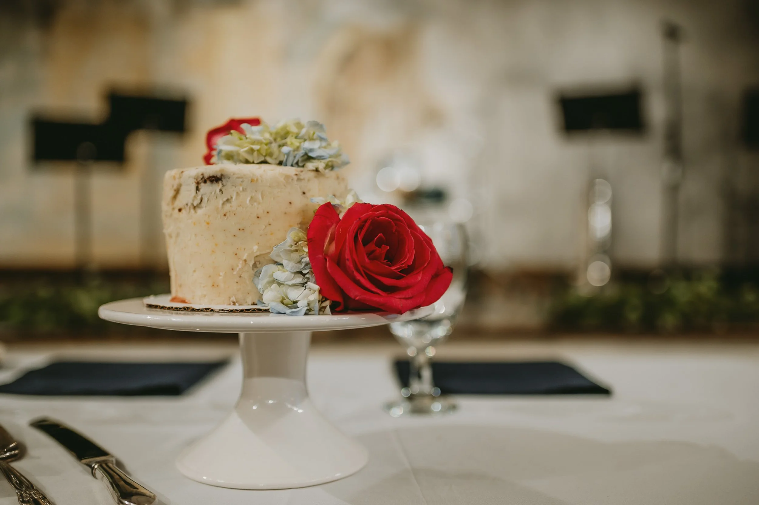 Small cake with white frosting and red roses on a pedestal stand, set on a table with cutlery and blurred background.