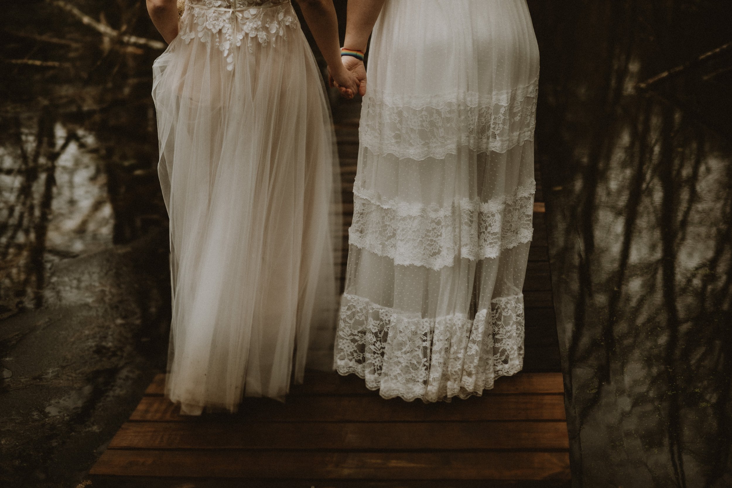 Two people wearing lace dresses, holding hands, standing outside on a wooden path.