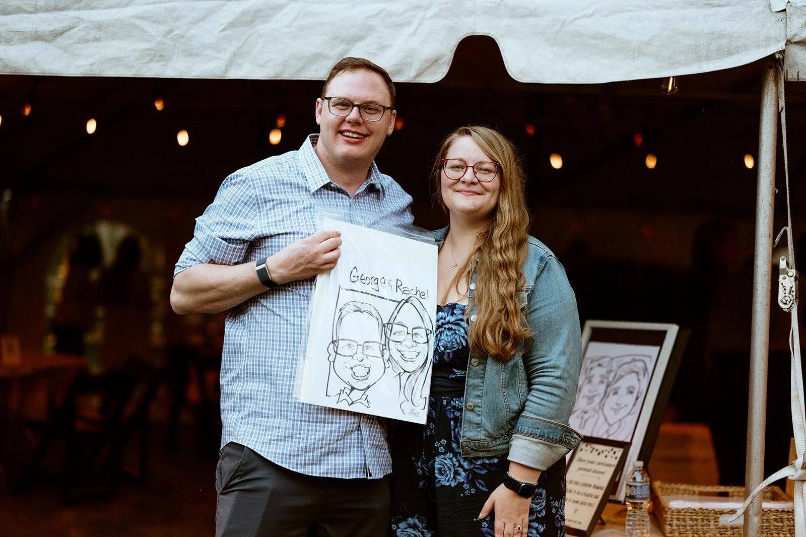 Two people smiling under a tent holding a caricature drawing of themselves.