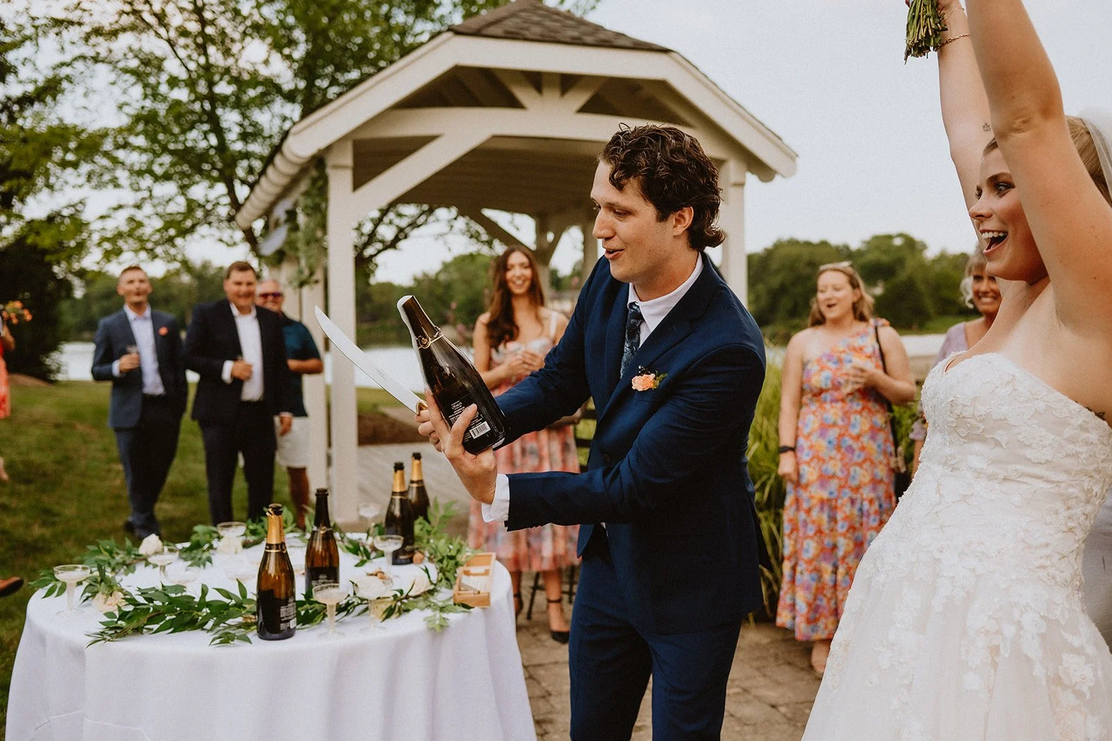A groom in a blue suit prepares to saber a champagne bottle at an outdoor wedding reception. The bride, in a white gown, raises her arm in excitement. Guests in formal attire watch and celebrate under a gazebo.