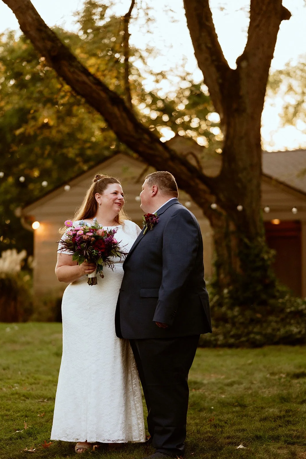A couple dressed in wedding attire is standing outdoors, with the bride holding a bouquet of flowers and the groom in a suit. They are gazing at each other lovingly in a garden setting, with trees and a building in the background.