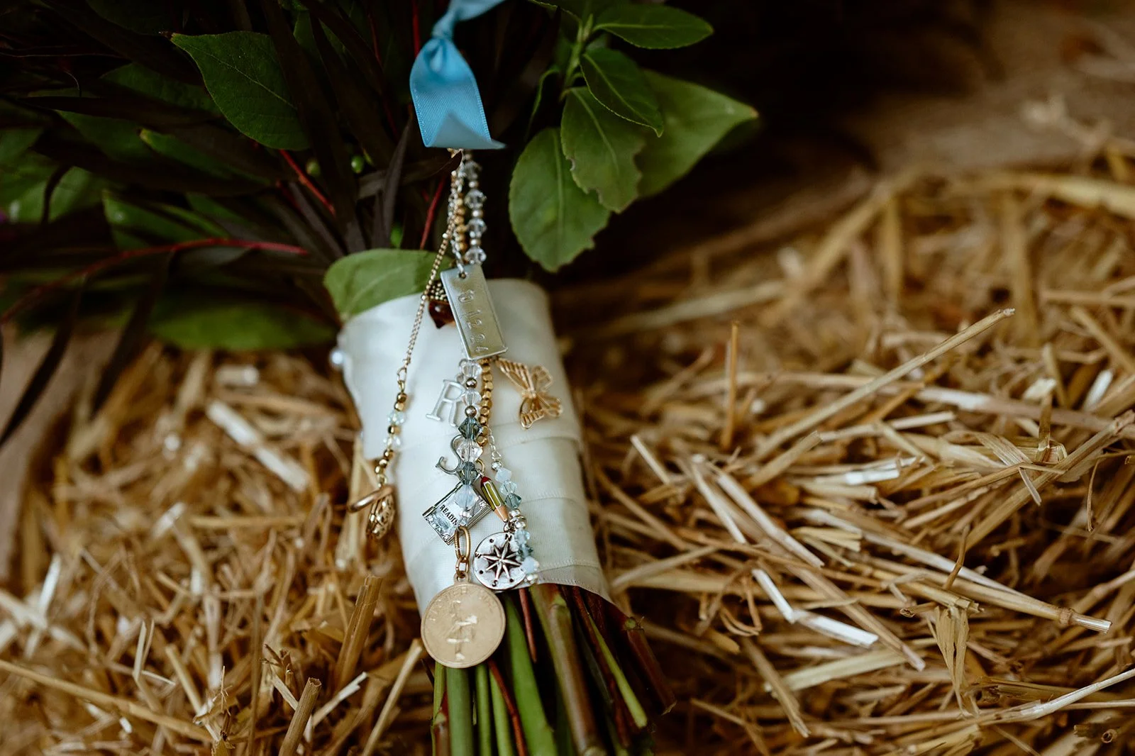 Close-up of a bouquet wrapped with white ribbon, adorned with various small charms and a gold chain, resting on a bed of straw.