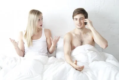 Man using his phone in bed while his wife expresses frustration